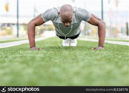 Fitness black man exercising push ups listening to music with headphones. Male model cross-training in urban background. African guy in his twenties doing workout outdoors in the street.