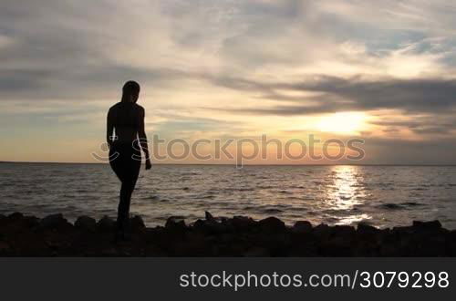 Fitness girl enjoying seascape on seaside with arms raised up towards the sky in glow of beautiful sunset. Back view of female silhouette proud of reaching her goals, achieving success with arms raised. Slow motion. Stabilized shot.