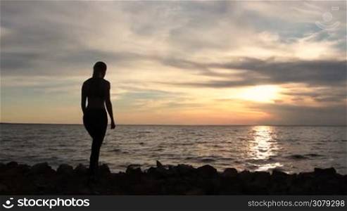 Fitness girl enjoying seascape on seaside with arms raised up towards the sky in glow of beautiful sunset. Back view of female silhouette proud of reaching her goals, achieving success with arms raised. Slow motion. Stabilized shot.