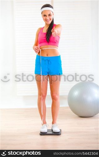 Fitness girl standing on weight scale and measuring her waist at home&#xA;