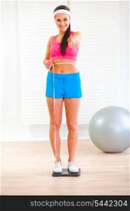 Fitness girl standing on weight scale and measuring her waist at home&#xA;