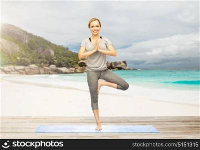 fitness, healthy lifestyle and people concept - woman doing yoga in tree pose on mat over exotic tropical beach background. woman doing yoga in tree pose outdoors on beach