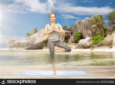 fitness, healthy lifestyle and people concept - woman doing yoga in tree pose on mat over exotic tropical beach background. woman doing yoga in tree pose outdoors on beach
