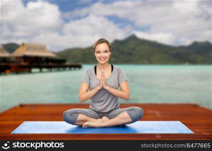 fitness, meditation and healthy lifestyle concept - happy smiling woman making yoga lotus pose on wooden pier over island beach and bungalow background. woman making yoga in and meditating lotus pose. woman making yoga in and meditating lotus pose