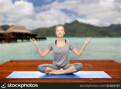 fitness, meditation and healthy lifestyle concept - happy woman making yoga lotus pose on wooden pier over island beach and bungalow background. woman making yoga in and meditating lotus pose