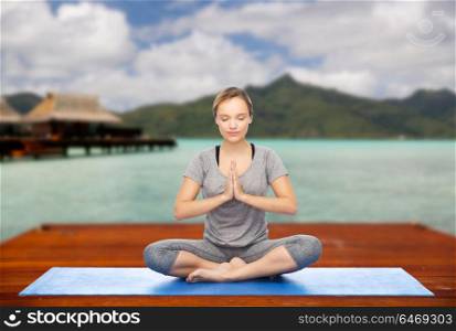 fitness, meditation and healthy lifestyle concept - happy woman making yoga lotus pose on wooden pier over island beach and bungalow background. woman making yoga in and meditating lotus pose