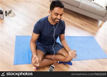 fitness, meditation and healthy lifestyle concept - indian man in earphones listening to music on smartphone and meditating in lotus pose at home. indian man meditating in lotus pose at home