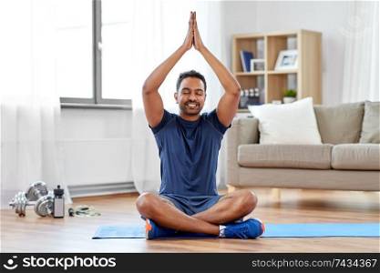 fitness, meditation and healthy lifestyle concept - indian man meditating in lotus pose on exercise mat at home. indian man meditating in lotus pose at home