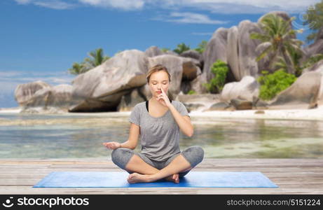 fitness, meditation and healthy lifestyle concept - woman doing yoga breathing exercise in lotus pose on mat over exotic tropical beach background. woman doing yoga breathing exercise on beach