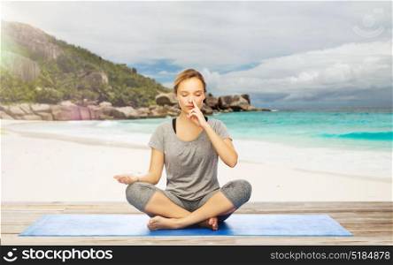 fitness, meditation and healthy lifestyle concept - woman doing yoga breathing exercise in lotus pose on mat over exotic tropical beach background. woman doing yoga breathing exercise on beach