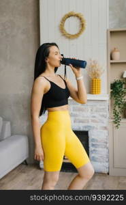 Fitness model in yellow leggings drinks water from a bottle while doing online lessons at home