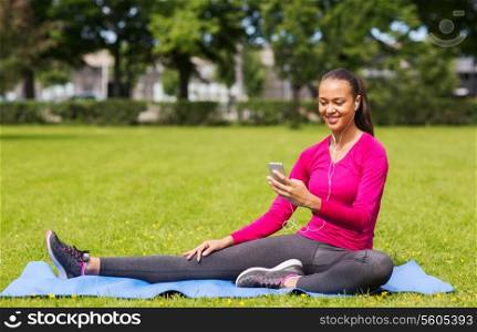 fitness, park, technology and sport concept - smiling african american woman with smartphone and earphones sitting on mat outdoors