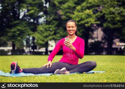 fitness, park, technology and sport concept - smiling african american woman with smartphone and earphones sitting on mat outdoors