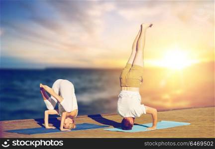 fitness, people and healthy lifestyle concept - couple doing yoga headstand outdoors on wooden pier over sea background. couple doing yoga headstand outdoors