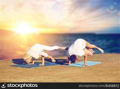 fitness, people and healthy lifestyle concept - couple doing yoga side crane pose on mat outdoors on wooden pier over sea background. couple doing yoga side crane pose outdoors
