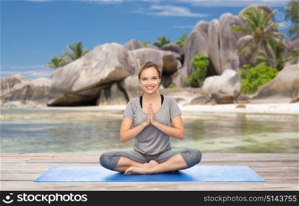 fitness, people and healthy lifestyle concept - happy smiling woman doing yoga meditation in lotus pose on mat over exotic tropical beach background. woman doing yoga meditation in lotus pose on beach