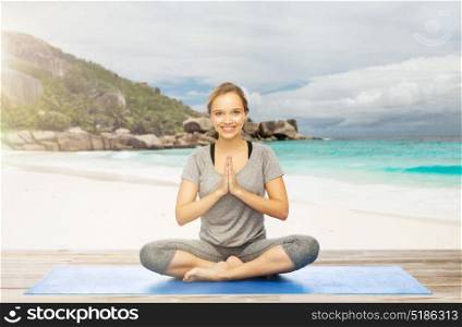 fitness, people and healthy lifestyle concept - happy smiling woman doing yoga meditation in lotus pose on mat over exotic tropical beach background. woman doing yoga meditation in lotus pose on beach
