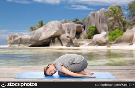 fitness, people and healthy lifestyle concept - happy woman doing yoga in child pose on mat over exotic tropical beach background. happy woman doing yoga in child pose on beach