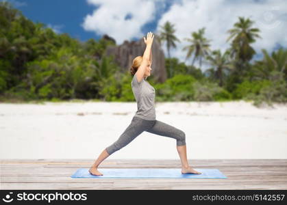 fitness, people and healthy lifestyle concept - happy woman doing yoga warrior pose on mat over exotic tropical beach background. happy woman doing yoga warrior pose on beach