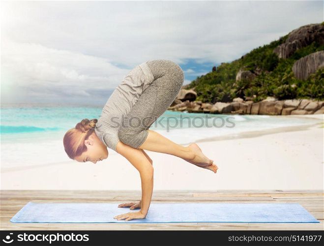 fitness, people and healthy lifestyle concept - woman doing yoga in crane pose on mat over exotic tropical beach background. woman doing yoga in crane pose on beach