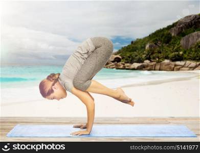 fitness, people and healthy lifestyle concept - woman doing yoga in crane pose on mat over exotic tropical beach background. woman doing yoga in crane pose on beach