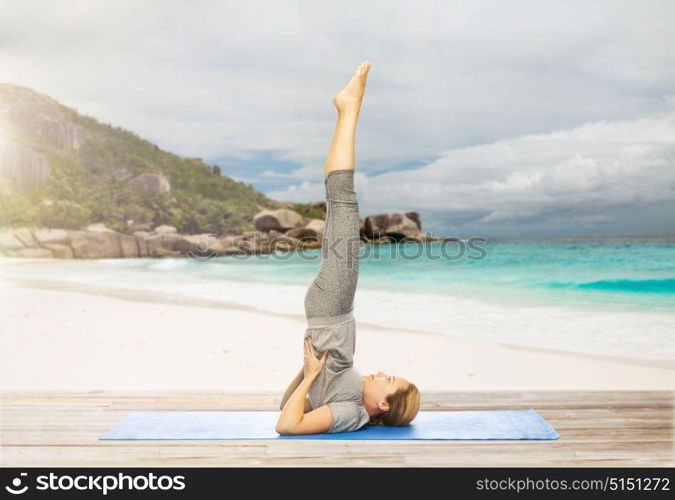 fitness, people and healthy lifestyle concept - woman making yoga in shoulderstand pose on mat over exotic tropical beach background. woman making yoga in shoulderstand pose on beach