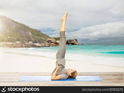 fitness, people and healthy lifestyle concept - woman making yoga in shoulderstand pose on mat over exotic tropical beach background. woman making yoga in shoulderstand pose on beach