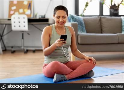 fitness, people and technology concept - happy smiling african american woman with earphones and smartphone doing sports at home. woman with earphones and smartphone doing sports