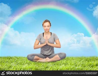fitness, people, harmony and healthy lifestyle concept - woman making yoga meditation in lotus pose over grass and rainbow in blue sky background