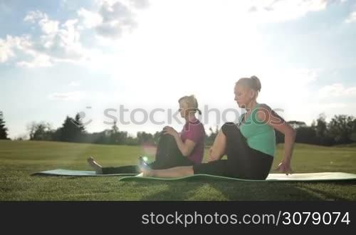 Fitness senior blonde women practicing yoga pose, doing pilates leg stretches exercises while sitting on exercise mats in park over amazing landscape background. Dolly shot. Adult fit females exercising abductor hip flexor muscle in buttocks outdoors