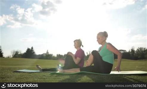 Fitness senior blonde women practicing yoga pose, doing pilates leg stretches exercises while sitting on exercise mats in park over amazing landscape background. Dolly shot. Adult fit females exercising abductor hip flexor muscle in buttocks outdoors