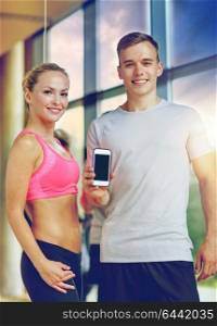 fitness, sport, advertising, technology and diet concept - smiling young woman and personal trainer with smartphone blank screen in gym. smiling young woman with personal trainer in gym