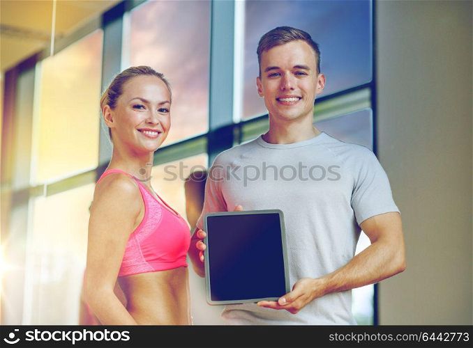 fitness, sport, advertising, technology and diet concept - smiling young woman and personal trainer with tablet pc blank screen in gym. smiling young woman with personal trainer in gym