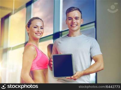 fitness, sport, advertising, technology and diet concept - smiling young woman and personal trainer with tablet pc blank screen in gym. smiling young woman with personal trainer in gym