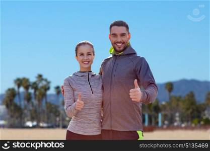 fitness, sport and gesture concept - smiling couple outdoors showing thumbs up over venice beach background in california. smiling couple in sport clothes showing thumbs up. smiling couple in sport clothes showing thumbs up