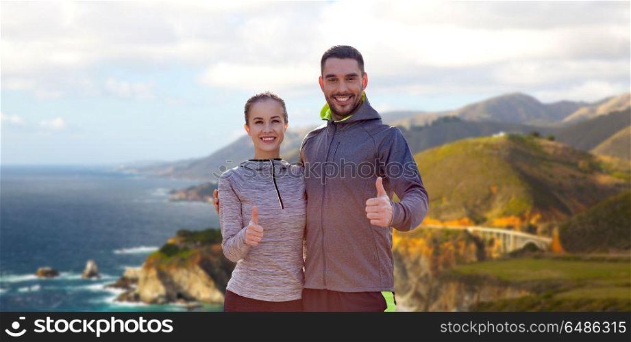 fitness, sport and gesture concept - smiling couple outdoors showing thumbs up over bixby creek bridge on big sur coast of california background. smiling couple in sport clothes showing thumbs up. smiling couple in sport clothes showing thumbs up