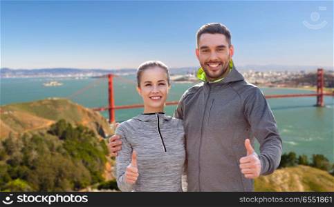 fitness, sport and gesture concept - smiling couple outdoors showing thumbs up over golden gate bridge in san francisco bay background. smiling couple in sport clothes showing thumbs up. smiling couple in sport clothes showing thumbs up