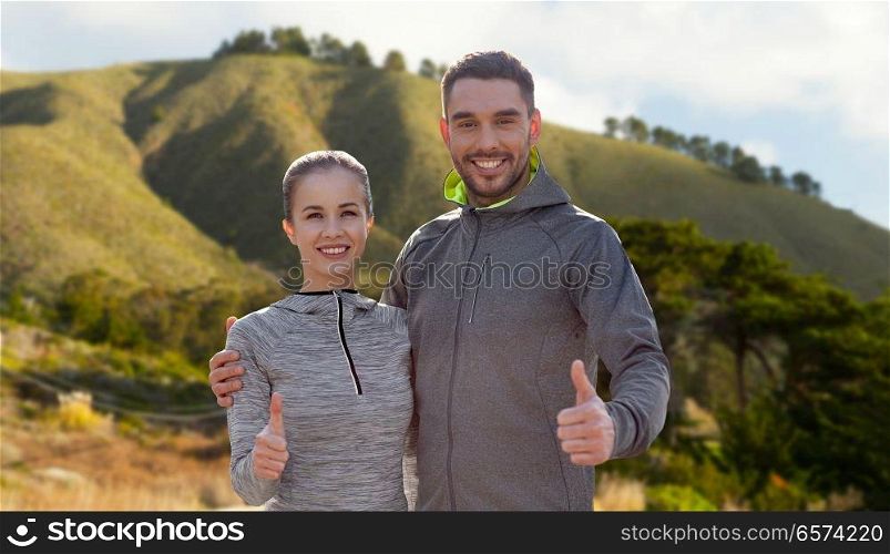 fitness, sport and gesture concept - smiling couple outdoors showing thumbs up over big sur hills background in california. smiling couple in sport clothes showing thumbs up