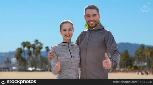 fitness, sport and gesture concept - smiling couple outdoors showing thumbs up over venice beach background in california. smiling couple in sport clothes showing thumbs up. smiling couple in sport clothes showing thumbs up
