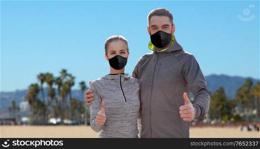 fitness, sport and health concept - couple wearing face protective black reusable masks for protection from virus disease showing thumbs up over venice beach background in california. couple in masks and sportswear showing thumbs up