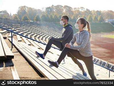 fitness, sport and health concept - couple wearing face protective medical mask for protection from virus disease stretching leg on stands of stadium. couple in masks stretching leg on stadium