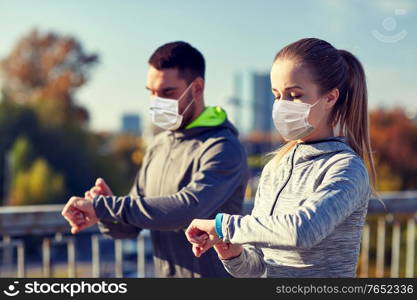 fitness, sport and health concept - couple wearing face protective medical mask for protection from virus disease with heart-rate watch doing sports over city highway bridge. couple in masks running over city highway bridge