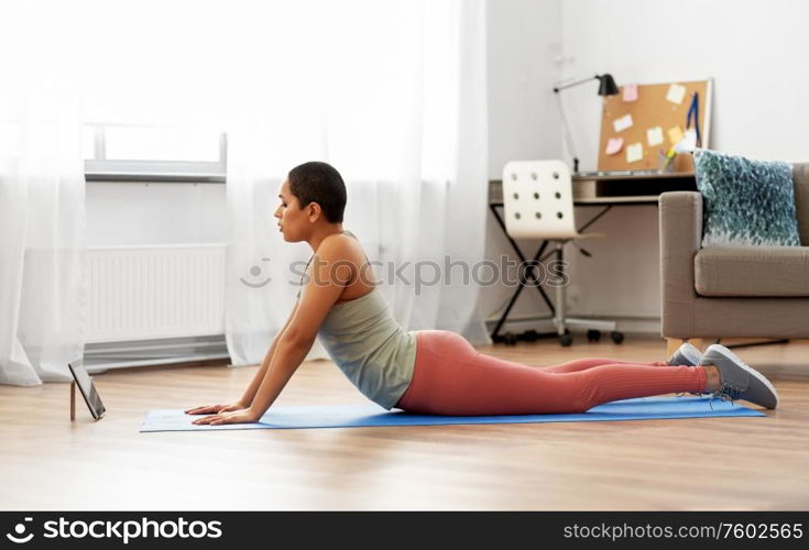 fitness, sport and healthy lifestyle concept - african american woman with tablet pc computer doing yoga cobra pose at home. woman with tablet pc doing yoga cobra pose at home