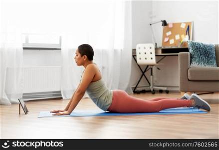 fitness, sport and healthy lifestyle concept - african american woman with tablet pc computer doing yoga cobra pose at home. woman with tablet pc doing yoga cobra pose at home