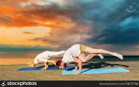 fitness, sport and healthy lifestyle concept - couple making yoga exercises outdoors on sea pier over sunset background. couple making yoga exercises outdoors
