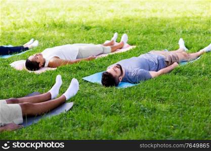 fitness, sport and healthy lifestyle concept - group of happy people doing yoga corpse pose or savasana at summer park. group of people doing yoga at summer park
