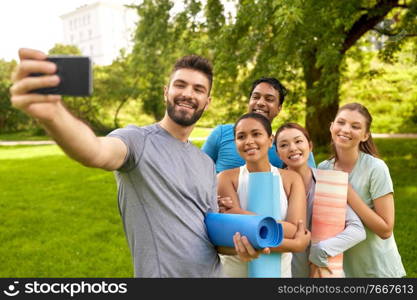 fitness, sport and healthy lifestyle concept - group of happy people with yoga mats taking selfie with smartphone at park. people with yoga mats taking selfie at park