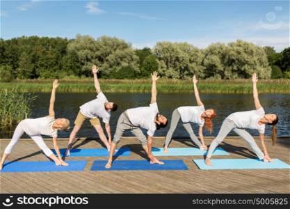 fitness, sport, and healthy lifestyle concept - group of people making left triangle pose on mat outdoors on river or lake berth. people making yoga in left triangle pose outdoors