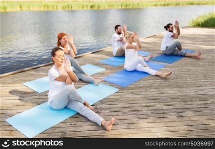 fitness, sport and healthy lifestyle concept - group of people making yoga exercise and meditating outdoors on river or lake berth. people making yoga and meditating outdoors