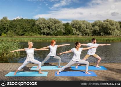 fitness, sport, and healthy lifestyle concept - group of people making yoga in warrior pose on river or lake berth. group of people making yoga exercises outdoors